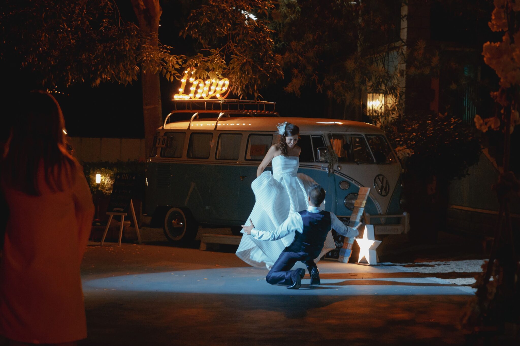 Novios bailando su primer baile de boda de noche frente a una furgoneta clásica vintage iluminada con letreros de luces.